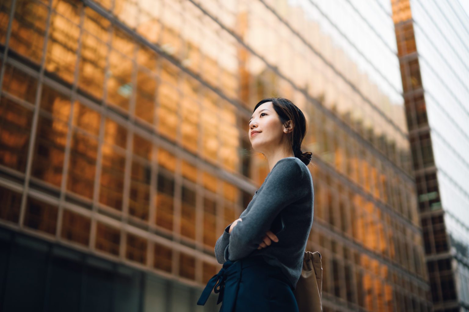 Low angle portrait of confident and successful young Asian businesswoman standing against contemporary corporate skyscrapers in financial district. Female leadership and determined to success